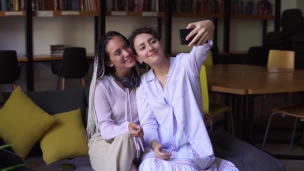 Unge kvinnelige studenter sitter på sofaen i et bibliotek omgitt av bøker. Jenta holder sin smartpone og tar selfie med sin venn fra universitetet med stilige dreadlocks og hodetelefoner på halsen. – stockvideo