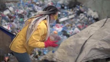 Sarı ceketli ve şeffaf koruma gözlüklü kadın çalışan modern geri dönüşüm fabrikasında kullanılmış plastik şişeleri ayırıyor. Geri dönüşüm fabrikasında büyük çuvallardan şişe fırlatmak.