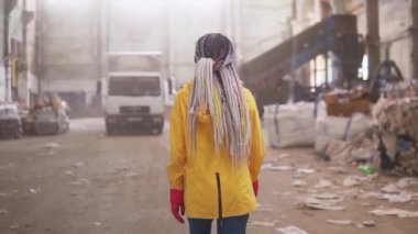 Koruyucu iş kıyafetleri içindeki bir kızın ender görüntülerini izliyoruz. Atık geri dönüşüm fabrikasının önünden geçiyor, geniş bir alanda kullanılmış şişeler, farklı atıklar ve arka planda beyaz kamyonlar var.