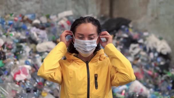 Eine junge Arbeiterin setzt sich eine Maske und eine Schutzbrille auf. Mädchen in gelber Jacke setzt Gesichtsmaske auf und bereitet sich auf den Arbeitstag in der Recyclingfabrik vor. Stapel gebrauchter Plastikflaschen auf Hintergrund — Stockvideo