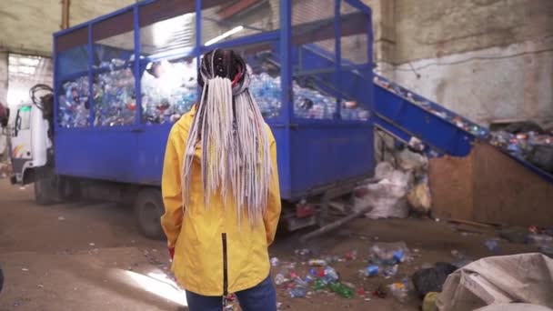 Baksida bilder av en kvinna med dreadlocks bär gul jacka titta på processen på avfall, återvinning fabrik. Stor lastbil med bur av begagnade plastflaskor för återvinning — Stockvideo