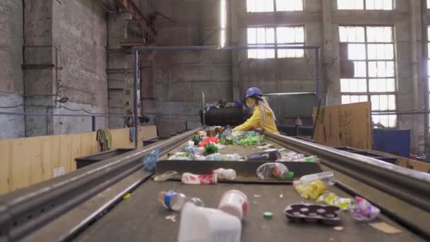 Vrouw-vrijwilliger in geel en transparant beschermende bril, harde hoed en masker sorteren gebruikte plastic flessen bij recyclingbedrijf. Aparte flessen op de lijn, het verwijderen van toppen en knijpen ze.Low hoek — Stockvideo