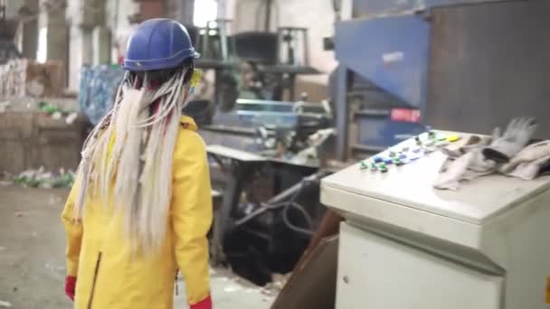 Mujer trabajadora con rastas en gafas protectoras amarillas y transparentes, gorro duro y máscara trabajando con equipos en planta de reciclaje. Máquina de prensa con botellas de plástico usadas y diferentes plásticos — Vídeo de stock