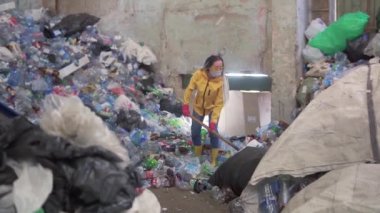 Sarı çizmeli, koruyucu maskeli, sarı çizmeli bir kız plastik geri dönüşüm fabrikasında büyük bir ördek küreğiyle şişe kullanıyordu. Arka planda kocaman bir şişe yığını var. Yavaş çekim