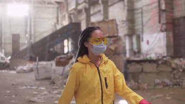 İş kıyafetleri içinde sarı ceketli, gözlüklü ve maskesiyle geri dönüşüm fabrikasında yürüyen kendine güvenen bir kız. Arkaplanda geniş, harabe alanı