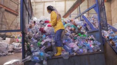 Sarı ceketli ve eldiven kepçeli genç bir kadının geri dönüşüm fabrikasında kullanılan plastik şişelerle çekilmiş görüntüleri. Araba kasasının içinde kocaman bir şişe yığını var.