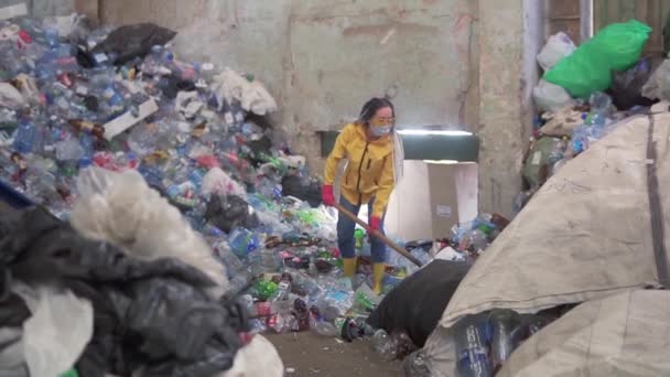 Dívka s dredy, ve žlutých botách v ochranné masce, nabírá láhve s velkou kachní lopatou v továrně na recyklaci plastů. Obrovská hromada lahví na pozadí. Zpomalený pohyb — Stock video