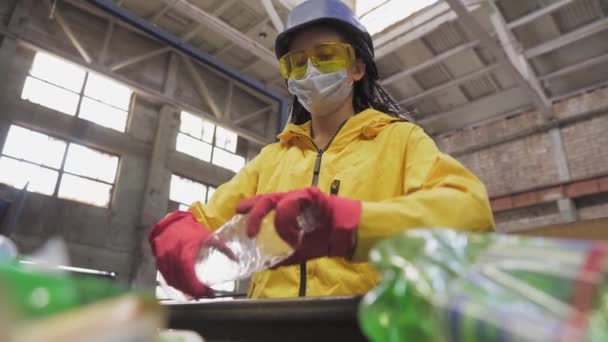 Volontaire-femme en veste jaune et lunettes de protection transparentes, casque et masque de tri des bouteilles en plastique usagées à l'usine de recyclage. Séparer les bouteilles sur la ligne, enlever les dessus et les presser — Video