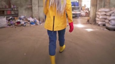Koruyucu iş kıyafetleri içindeki bir kızın ender görüntülerini izliyoruz. Atık geri dönüşüm fabrikasının önünden geçiyor, geniş bir alanda kullanılmış şişeler, farklı atıklar ve beyaz kamyonlar var. Kapat.
