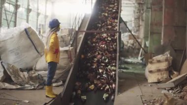 Sarı ve şeffaf koruyucu gözlüklü kadın işçi, koruyucu şapka ve maske, kullanılmış plastik şişelerle dolu taşıyıcıyı kaldırırken izliyor. Otomatik işlemin geri dönüşüm fabrikasındaki görüntüsü