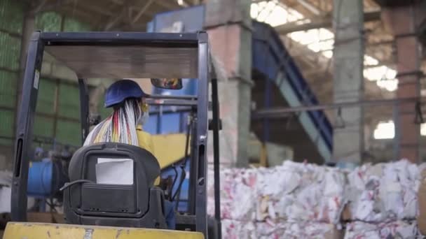 Vykládání odpadního papíru ve skladovém elektromobilu. Dělnice v klobouku a žluté bundě sedící ve stroji. Obrovské zásoby lisovaného kartonu. Žena řidič sedí zezadu a ukazuje na — Stock video