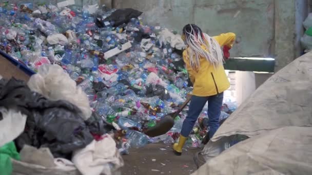 Een meisje met dreadlocks, in gele laarzen, gebruikte flessen met een grote eendenschop in een plastic recyclingfabriek. Enorme stapel flessen op de achtergrond — Stockvideo