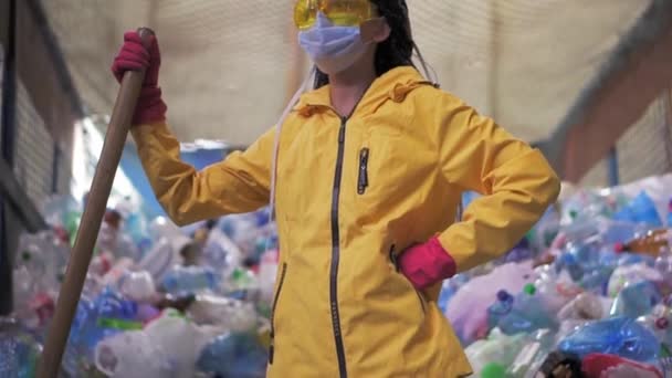 노란 자켓과 마스크를 입고 커다란 오리 삽을 들고 플라스틱 재활용 공장에 서 있는 소녀의 모습. 뒤에 커다란 병들이 쌓여 있습니다. 낮은 각도 전망, 고개를 돌리고 — 비디오