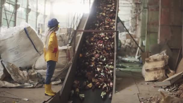 Sarı ve şeffaf koruyucu gözlüklü kadın işçi, koruyucu şapka ve maske, kullanılmış plastik şişelerle dolu taşıyıcıyı kaldırırken izliyor. Otomatik işlemin geri dönüşüm fabrikasındaki görüntüsü — Stok video