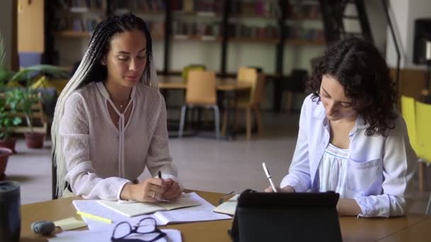 Porträt zweier attraktiver Frauen, die in einer Bibliothek mit Bücherregalen im Hintergrund ein Gespräch führen. Mit Tablet und diversen Papieren am Holztisch sitzend. Dreadlocks Frau erklärt etw. — Stockvideo