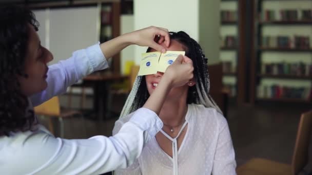 Two caucasian women making fun during their preparation for exams. Brunette girl sticks yellow papers with painted eyes on her female friends eyes — Stock Video
