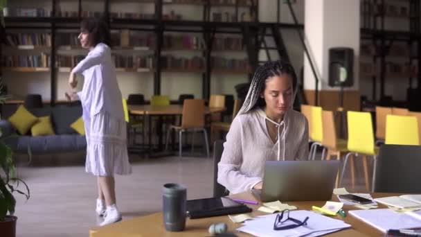 Morena feliz em roupas casuais brancas em fones de ouvido dança engraçada no fundo, enquanto sua amiga se concentrou em estudar, sentada à mesa funciona com laptop. Movimento lento — Vídeo de Stock