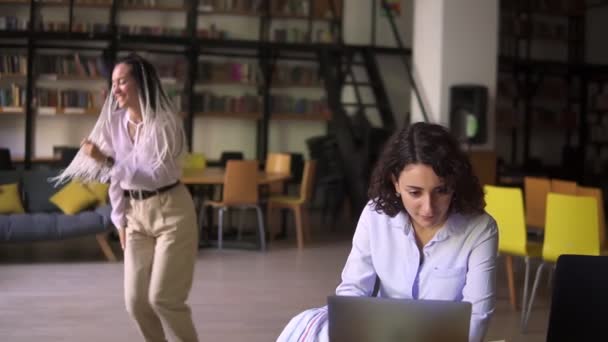 Biblioteca pública aberta. Happy dreadlocks menina bonita em blusa branca e bege calças engraçadas dançando no fundo, enquanto sua amiga se concentrou em estudar, sentado à mesa funciona com laptop — Vídeo de Stock