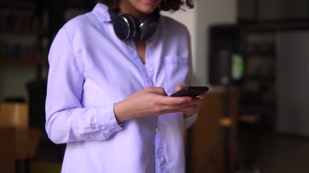 Corte imagens de mulher morena andando usando blusa branca, olhando na tela do smartphone em suas mãos e digitando, fones de ouvido no pescoço. Caminhando pela moderna biblioteca pública concentrada com seu gadget — Vídeo de Stock