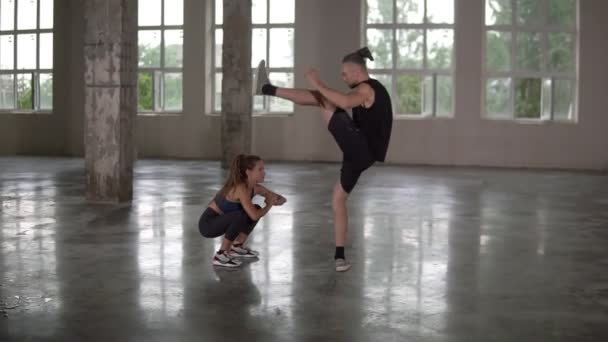 Hermosa mujer joven con rastas haciendo sentadillas ejercicio con el hombre de la pareja levantando las piernas bajo su cabeza sincrónicamente. Estudio loft fitness con grandes ventanas en el fondo — Vídeos de Stock