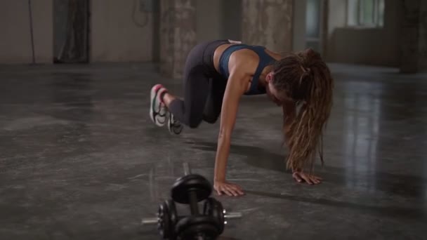 Jonge sportieve stijlvolle vrouw trainen haar lichaam training haar spieren indoor in lege studio. Atletische mooie stijlvolle vrouw met lange dreadlocks doet hardloopplank oefening — Stockvideo