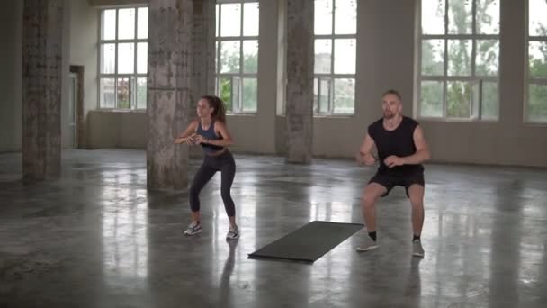 Ung man och kvinna med dreadlocks tränar på tom studio, gör synkron knäböj tillsammans, stark och frisk, upprätthålla sin aktiva livsstil, slow motion — Stockvideo