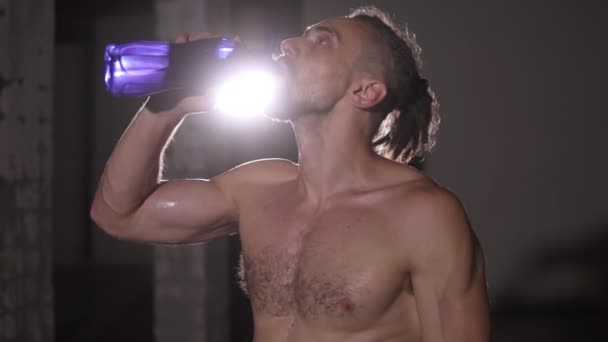 Culturismo hombre con rastas retrato de pie delante de la cámara sin camisa beber sensualmente agua de la botella azul en luces foco de fondo. Movimiento lento — Vídeo de stock