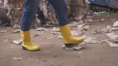 Koruyucu iş kıyafetleri içinde tanınmayan bir kadın. Atık geri dönüşüm fabrikasının yanından geçen sarı çizmeler kullanılmış şişe ve çöplerle dolu büyük bir alan. Görüntüleri kapat