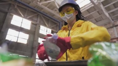 Sarı ve şeffaf koruma gözlüğü takan kadın gönüllü, geri dönüşüm fabrikasında kullanılmış plastik şişeleri ayırıyor. Çizgide ayrı şişeler var, üstlerini çıkarıp sıkıyorlar. Düşük açı