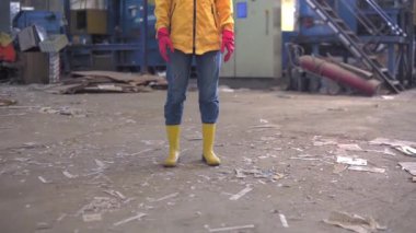 Koruyucu iş kıyafetleri içinde rastalı bir genç kız. Sarı ceket, gözlük, şapka ve maske. Geri dönüşüm fabrikasının arkasında duruyor. Özel ekipman, arka planda makine