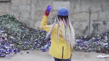 Sert şapkalı kadın kırık cam yığınına karşı duruyor, duvarın yanında kullanılmış şişeler var. Sarı ceketli kız geri dönüşüm için eski cam şişeleri çarpıyor. Nadir bir manzara. Yavaş çekim