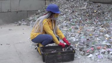 Sert şapkalı kadın kırık cam yığınından kırık olmayan cam şişeleri topluyor, duvarın yanında kullanılmış şişeler. Sarı ceketli kız daha ileri gitmek için eski cam şişeleri topluyor.