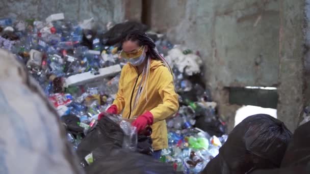 Wanita berjaket kuning, kacamata pelindung dan sarung tangan memilah botol plastik dari kantong hitam. Tumpukan besar botol plastik bekas di latar belakang — Stok Video