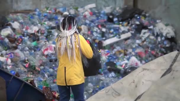 Stylowa kobieta w żółtej kurtce, okulary ochronne i rękawice sortujące plastikowe butelki z czarnych toreb. Ogromny stos używanych plastikowych butelek różnych kolorów na tle — Wideo stockowe