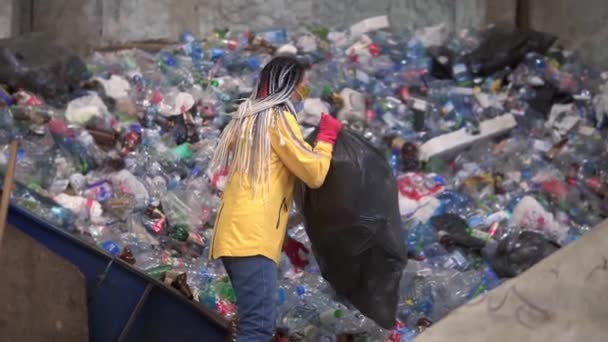 Kvinnlig arbetare i gul jacka och skyddsglasögon packa upp svart suger med begagnade plastflaskor på modern återvinningsanläggning. Kasta flaskor från stor säck på hög i återvinningsfabrik — Stockvideo