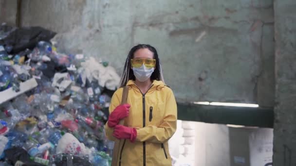 Portrét dívky s dredy, ve žluté bundě a masce, držící velkou kachní lopatu, stojící v továrně na recyklaci plastů. Obrovská hromada lahví na pozadí — Stock video