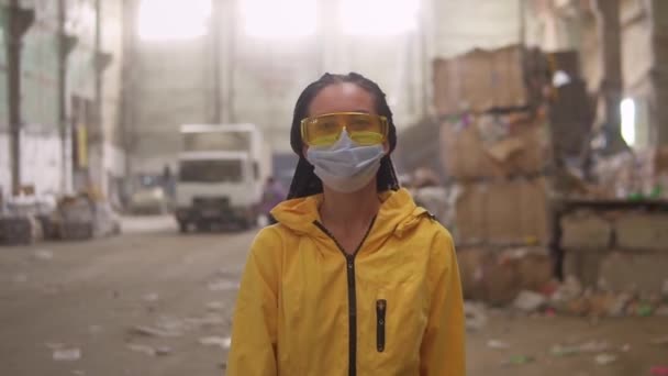 Une fille positive en vêtements de travail protecteurs - veste jaune, lunettes et masque marchant en recyclant l'usine et souriant. Boîtes de déchets, camion sur fond — Video