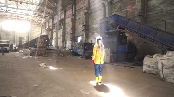 天井から自然光が射す中、空の廃棄物再資源化工場の真ん中に黄色のジャケットとブーツを着たドレッドロックの女性の映像 — ストック動画