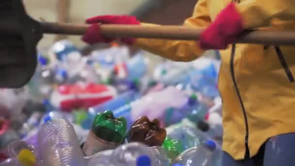 Nahaufnahme einer Person in gelber Jacke und Handschuhen, die mit einer großen Entenschaufel gebrauchte Flaschen in einer Kunststoff-Recyclingfabrik schaufelt. Riesiger Flaschenhaufen im Hintergrund — Stockvideo
