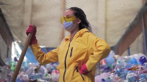 Portret dziewczyny z dredami, w żółtej kurtce i masce, trzymającej dużą łopatę z kaczki, stojącej w fabryce recyklingu tworzyw sztucznych. Ogromny stos butelek na tle. Widok z niskiego kąta — Wideo stockowe