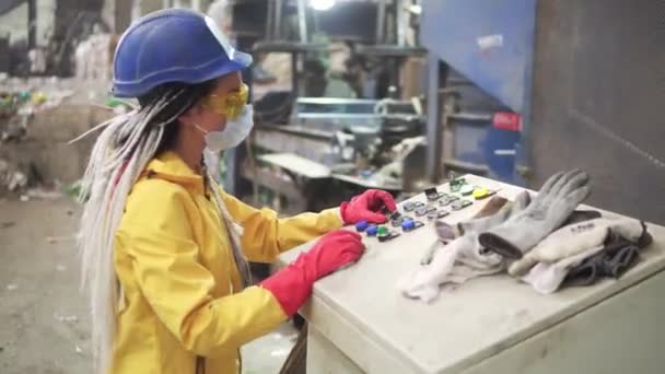 Mujer trabajadora en gafas protectoras amarillas y transparentes, gorro duro y máscara que trabaja con equipos en planta de reciclaje. Máquina de prensa con botellas de plástico usadas y diferente basura de plástico. Imágenes de — Vídeo de stock