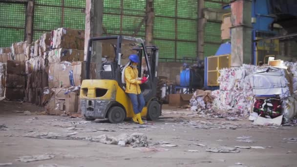Eine Arbeiterin mit Bollenhut und gelber Jacke steht, lehnt an einem Elektroauto und liest Arbeitstagebuch. Riesige Vorräte an gepresstem Karton. Volle Länge — Stockvideo
