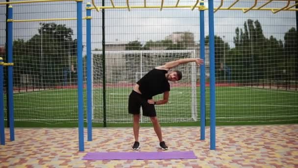 Full längd av stilig skäggig ung idrottsman på stadion uppvärmning, stretching, gör sidokurvor — Stockvideo