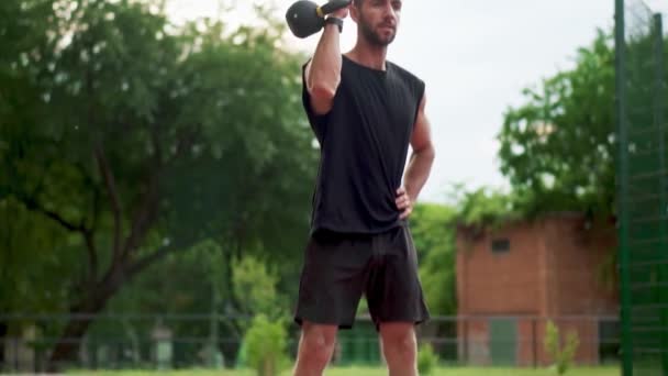 Un uomo barbuto in abbigliamento sportivo nero si allena con peso bob all'aperto vicino allo stadio in pista in slow mo — Video Stock