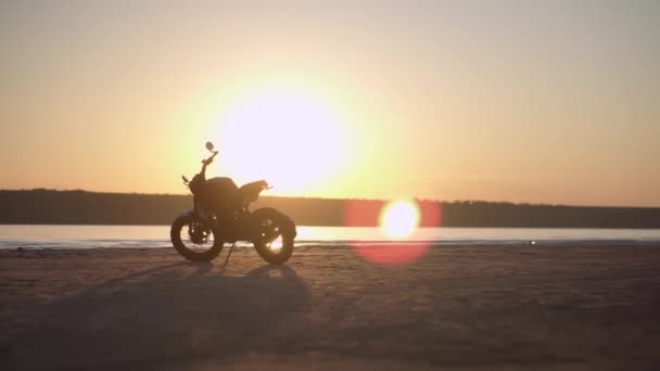 Egy motorkerékpár parkoló a földön, közel a vízhez naplementében, vízvonallal a háttérben. Alacsony látószög — Stock videók