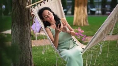 Esmer bir kadın açık havada hamakta dinleniyor, akıllı telefonuyla internette sörf yapıyor, ekrana tıklıyor.