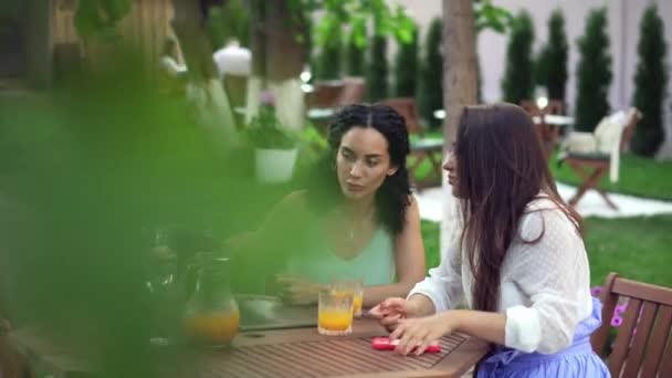 Две женщины сидят в кафе на открытом воздухе - разговаривают, делятся новостями, девочки показывают что-то на экране ноутбука и смартфона — стоковое видео
