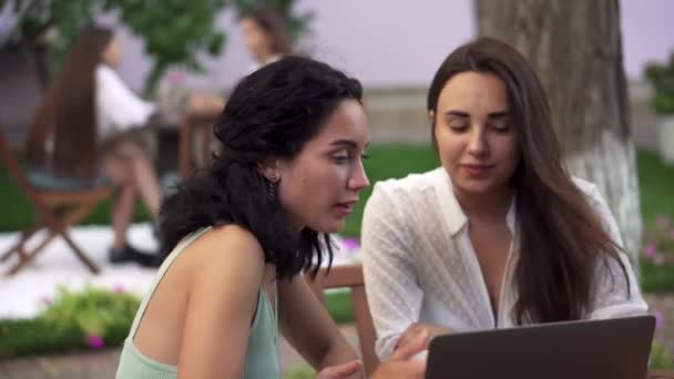 Две женщины сидят в кафе на открытом воздухе - разговаривают, делятся новостями, брюнетка девушка показывает что-то на экране ноутбука, жесты — стоковое видео