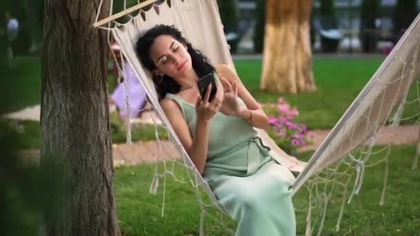 Mujer morena relajándose en una hamaca al aire libre, navegar por Internet en su teléfono inteligente, clics en la pantalla — Vídeos de Stock