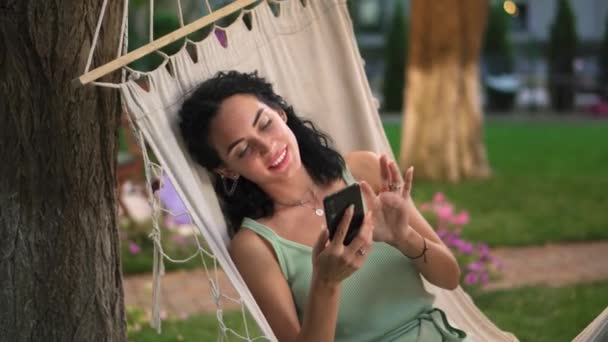 Retrato de una mujer relajándose en una hamaca al aire libre, navegando por Internet en su teléfono inteligente, clics en la pantalla y sonriendo — Vídeos de Stock